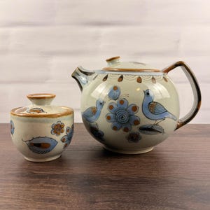 May include: A ceramic teapot and a matching lidded sugar bowl featuring a hand-painted design of blue birds, orange and blue flowers, and brown leaf motifs on a cream-colored background with brown glazed rims and a brown handle.