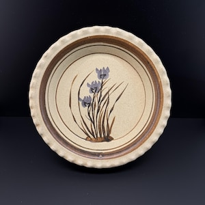 May include: A round ceramic pie dish with a fluted edge. The plate has a beige interior with a brown rim and a floral design of purple irises and green stems. Suitable for baking and serving, it measures approximately 25 cm in diameter.