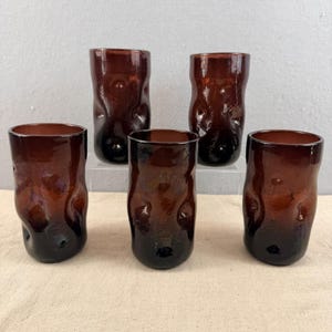 Puede incluir: Cinco vasos de vidrio teñidos de marrón con un diseño texturizado único. Los vasos tienen una base oscura que se desvanece a un marrón más claro hacia la parte superior. Los vasos están dispuestos sobre una superficie neutra.