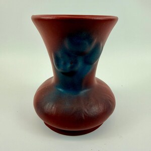 May include: A matte terracotta-colored ceramic vase with a wide, flared rim and a bulbous base. The surface features a smooth texture with a prominent, dark blue, cloud-like glaze pattern concentrated around the neck and upper body area.