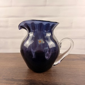 Pode incluir: Um jarro de vidro roxo escuro com borda ondulada e uma alça de vidro transparente. O jarro apresenta um corpo arredondado e um pequeno adesivo quadrado com o texto Blenko Handmade visível na superfície frontal do recipiente de vidro.