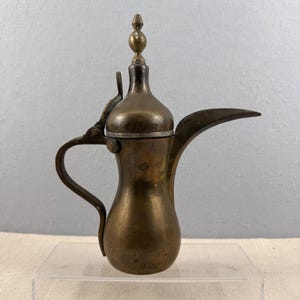 May include: A vintage brass coffee pot with a long spout and curved handle. The pot has a bulbous body and a decorative finial on the lid. The metal has a tarnished, aged appearance, suggesting it is an antique. The pot is displayed on a clear surface.