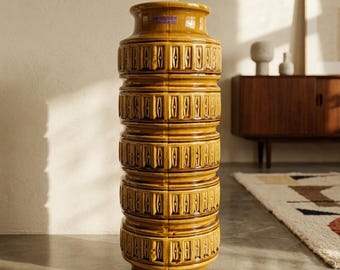 May include: A tall, cylindrical ceramic floor vase with a glossy mustard yellow glaze. The exterior features a repeating vertical geometric pattern of recessed rectangular panels with central diamond shapes. A small purple label reads Scheurich.