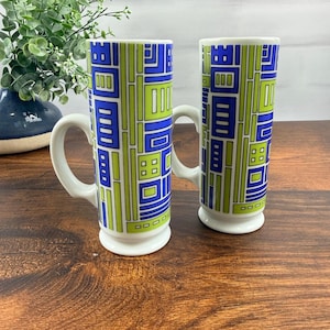 Puede incluir: Dos tazas altas de cerámica con un patrón geométrico de rectángulos y líneas en azul, verde lima y blanco. Cada taza cuenta con un asa blanca, un cuerpo cilíndrico y una base circular elevada, manteniendo un diseño gráfico uniforme.