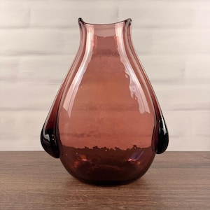 Pode incluir: Um vaso de vidro malva translúcido com um corpo arredondado em forma de lágrima e um pescoço largo e aberto. O vaso apresenta duas saliências de vidro de cor escura nos lados inferiores, conferindo-lhe uma silhueta com bordas curvas.
