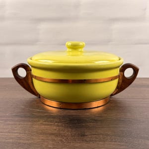 May include: A bright yellow ceramic casserole dish with a matching lid, featuring a smooth glossy finish. The bowl is encircled by a thin copper band and includes two dark brown wooden handles with circular cutouts on opposite sides.