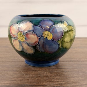 Peut inclure: Un bol rond en céramique avec une base bleu sarcelle foncé orné de motifs floraux peints à la main. Le design présente de grandes fleurs roses et violettes aux centres jaunes et des feuilles vertes, le tout avec un fini glacé et craquelé.