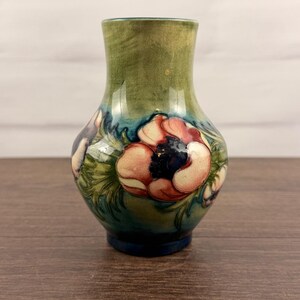 Puede incluir: Un jarrón de cerámica con cuerpo bulboso y cuello acampanado, que presenta un diseño pintado a mano de grandes flores de anémona rosas y blancas con centros oscuros, sobre un fondo mezclado de tonos verde musgo, azul cerceta y azul profundo.