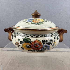 May include: Cream-colored ceramic casserole dish with lid, adorned with colorful floral patterns in orange, yellow, blue, and green. The dish has brown handles and a gold rim, perfect for serving. The lid is topped with a brown finial.