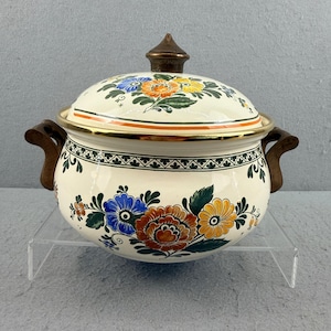 May include: Cream-colored enamelware pot with lid, adorned with floral patterns in blue, orange, and yellow. The pot features wooden handles and a bronze-colored knob on the lid. A gold band encircles the rim, adding a touch of elegance.