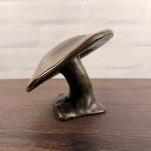 Peut inclure: Une sculpture de champignon en céramique avec un chapeau brun lisse et brillant et un pied aux tons métalliques sombres. La pièce présente un chapeau large et incurvé qui surplombe une base épaisse et solide, avec une finition polie.