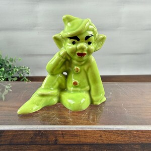 May include: A glossy lime green ceramic figurine of a seated elf with pointed ears, a cap, and two brown buttons on the chest. The figure has painted black eyebrows and red lips, depicted in a resting pose with one hand near the face.