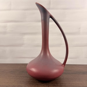 May include: A tall, slender ceramic pitcher with a matte, deep burgundy finish that transitions to a darker, near-black tone at the elongated spout. The vessel features a wide, rounded base and a single, curved handle that connects the neck to the body.