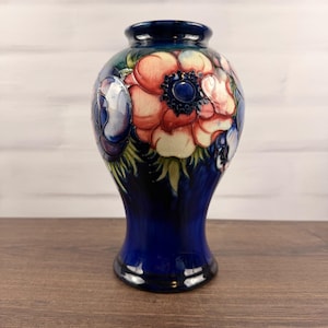 Puede incluir: Un jarrón de cerámica con una base de color azul cobalto profundo que se transforma en un patrón floral con grandes flores de anémona de color rosa claro y crema con centros azul oscuro, acentuado por hojas verdes y motivos florales azules.