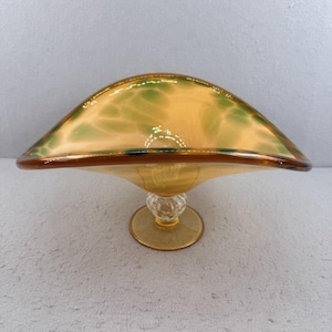 May include: A decorative glass bowl with a wide, curved rim. The bowl features a yellow and green mottled design with a brown rim and a clear, ornate base. The base is supported by a round, yellow foot.