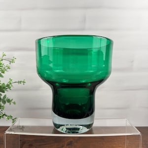 Pode incluir: Um vaso de vidro verde esmeralda profundo com uma abertura superior larga e alargada e uma base de vidro transparente espessa. O recipiente apresenta uma superfície lisa e polida com um corpo cilíndrico que se estreita ligeiramente.