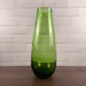 Peut inclure: Un vase haut et effilé en verre vert translucide. Le récipient présente une silhouette lisse et arrondie qui se rétrécit vers l'ouverture supérieure. La partie inférieure du corps en verre contient de nombreuses petites bulles d'air.