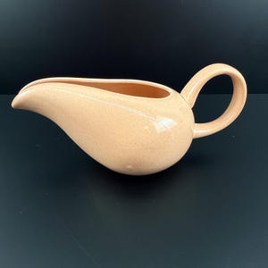 Puede incluir: Una salsera de cerámica color melocotón con cuerpo redondeado y pico curvo. La salsera tiene un asa y una textura moteada. El fondo es negro liso.