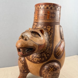 May include: A tan-colored ceramic vessel with a jaguar-like face and intricate brown patterns. The vessel has a cylindrical neck and a rounded body, with the jaguar's head forming the front. The design includes geometric shapes and stylized animal motifs.