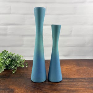 Op de afbeelding: Twee hoge, slanke kandelaars met een gladde, matte afwerking. De kandelaars hebben een kleurverloop dat overgaat van een licht hemelsblauw aan de bovenkant naar een dieper, donkerder blauw aan de basis. Beide objecten hebben een kegelvorm.