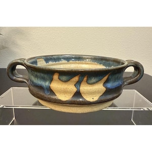 May include: A handcrafted ceramic bowl with two handles. The bowl has a blue and brown glaze with a distinctive design. The interior is a light beige colour. The bowl is suitable for serving soups, stews, or other dishes.