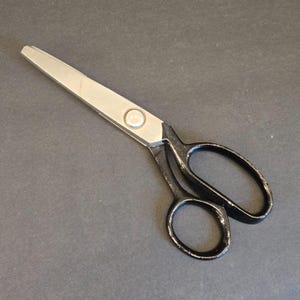 May include: A pair of scissors with a silver blade and black handles. The scissors are lying on a dark gray surface. The blades are connected with a silver rivet.