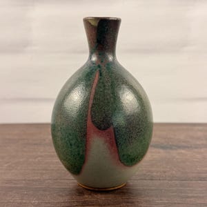 May include: A small ceramic bud vase with a rounded body and a narrow neck. The surface features a mottled glaze with deep forest green, dark brown, and muted mauve tones that blend together in a smooth, glossy finish across the entire vessel.
