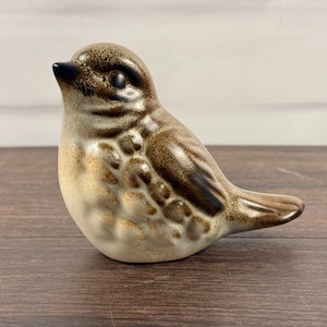 May include: A small ceramic bird figurine featuring a gradient of colours ranging from a dark brown head and wings to a lighter cream-coloured chest and belly. The surface has a smooth, glazed finish with detailed moulded feather textures.