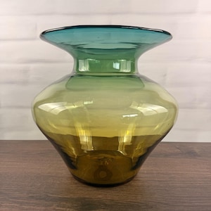 Puede incluir: Un florero de vidrio con un borde ancho y acampanado y un cuerpo redondeado. El vidrio muestra una transición de color degradada, comenzando con un azul verdoso profundo en la parte superior, desvaneciéndose a verde claro y terminando en tono ámbar oscuro.