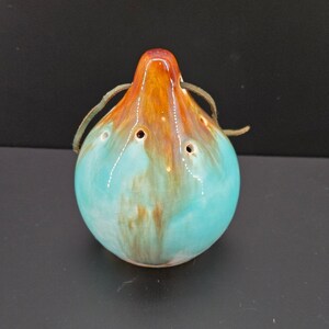 Puede incluir: Ocarina de cerámica en forma de lágrima, con cuerpo turquesa y detalles marrones y naranjas. La ocarina tiene agujeros para los dedos y un cordón de cuero para colgar.