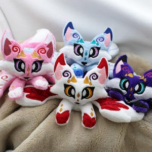 May include: Four plush fox toys with multiple tails, featuring embroidered facial details and swirling patterns. The foxes are colored in pink, light blue, white with red accents, and deep purple with white accents, all with large, expressive eyes.