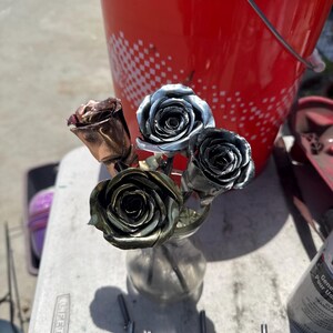May include: A collection of four metal roses in a small glass vase. The roses feature distinct metallic finishes, including copper, silver, and dark bronze tones. Each flower is crafted with layered petals, creating a realistic floral structure.