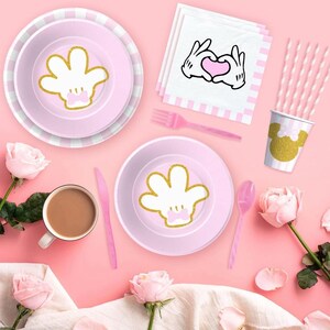 May include: A pink party table setting with plates, cutlery, and a cup featuring a gold glitter hand design. Pink and white striped straws and napkins with a hand-heart graphic are also present. The setting is decorated with pink roses.