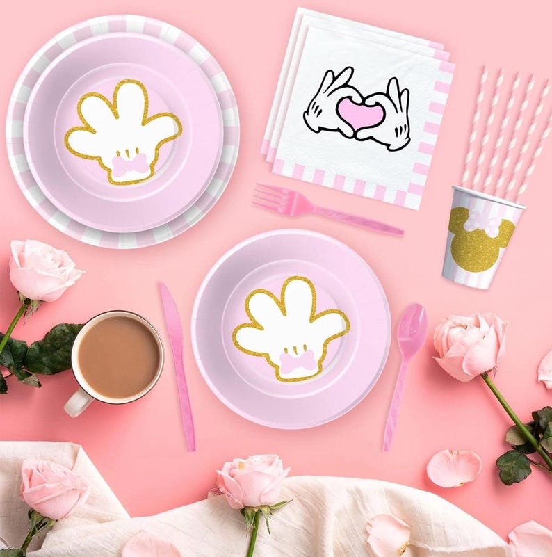 May include: A pink party table setting with plates, cutlery, and a cup featuring a gold glitter hand design. Pink and white striped straws and napkins with a hand-heart graphic are also present. The setting is decorated with pink roses.