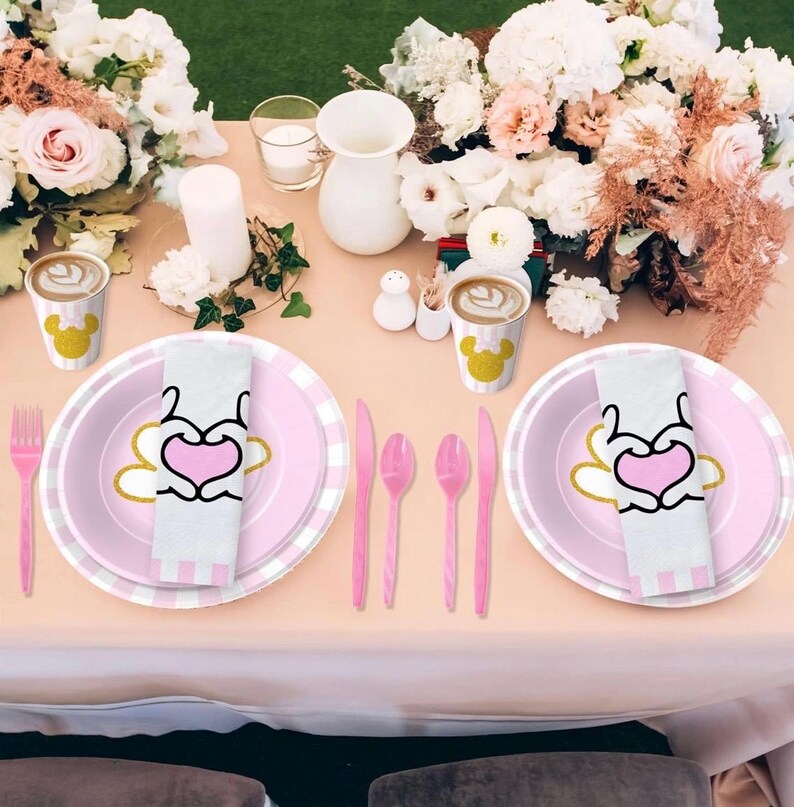 May include: A table setting with pink and white plates, napkins, and cutlery. The plates have a heart design with gold accents. Coffee cups with a yellow mouse head are also present, along with flowers and candles.