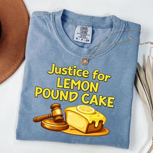 May include: A blue Comfort Colors t-shirt with the text "Justice for Lemon Pound Cake" in yellow. The graphic includes a gavel and a slice of lemon pound cake.