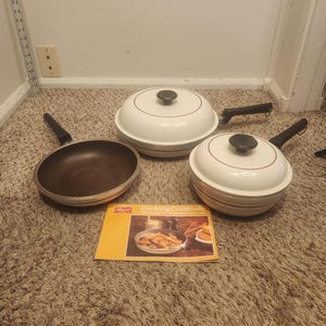 Puede incluir: Un juego de tres piezas de cocina blancas y negras que incluye una sartén abierta y dos ollas con tapa y mangos negros. Se incluye un folleto amarillo titulado Cooking Creatively. Una regla muestra una altura de 50 cm.