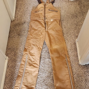 May include: Tan work overalls with brown straps and a front zip. The overalls feature multiple pockets and zips along the outer seams of the legs. The garment is made of a durable fabric.