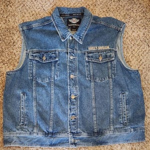 May include: A blue denim vest featuring a classic collar, a button-down front, and two chest pockets with buttoned flaps. The text HARLEY-DAVIDSON is embroidered in white above the left chest pocket, and a logo patch is visible on the inner neck label.