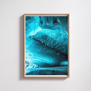 Op de afbeelding: Een ingelijste print met een abstracte weergave van een ijsgrot. Het interieur van de grot is verlicht met tinten turquoise en blauw, met een textuur en vloeiend uiterlijk. De print is gevat in een lichtgekleurde houten lijst.