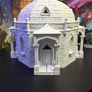 Puede incluir: Un modelo detallado en gris claro de un edificio abovedado, probablemente para juegos de mesa. La estructura presenta ventanas arqueadas, columnas decorativas y un exterior texturizado similar a ladrillos. El modelo se encuentra sobre cajas de juego de Dungeons & Dragons.