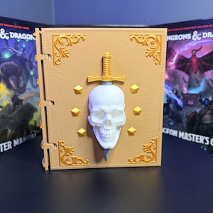 May include: A tan-colored book with gold accents, a white skull, and a sword. The book has a spine with hinges and is decorated with gold corner designs and diamond-shaped studs. The text "Dungeons & Dragons" is visible in the background.