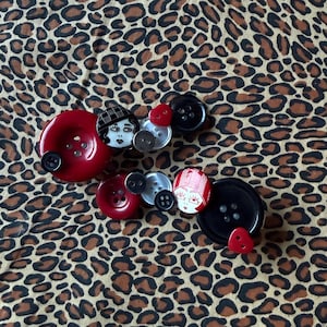 Pode incluir: Uma variedade de botões decorativos em vários tamanhos e cores, incluindo vermelho, preto e prata. Alguns botões apresentam rostos pintados. Os botões estão dispostos sobre um tecido com estampa de leopardo.