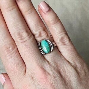 May include: A silver ring with an oval turquoise stone. The ring has a decorative silver band with a textured design. The turquoise stone is a vibrant shade of blue-green. The ring is shown on a person's finger.