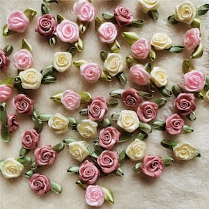 May include: A collection of small fabric ribbon roses in shades of light pink, dusty rose, and cream. Each individual rose features tightly wound satin petals and a small green fabric leaf attached to the base, scattered across the frame.