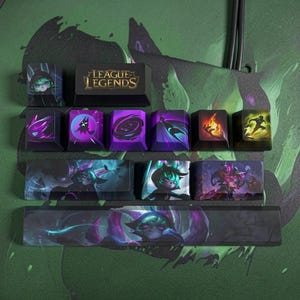 May include: A set of black mechanical keyboard keycaps featuring League of Legends branding and character graphics. The keys display purple, green, and orange illustrations of a hooded figure, magical effects, and flames on a dark background.