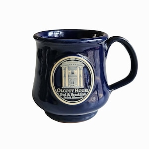 May include: A deep blue ceramic mug featuring a circular cream-colored emblem on the front. The emblem depicts a building with columns and the text "1904 Olcott House Bed & Breakfast Duluth, Minnesota" printed in dark blue lettering on the surface.