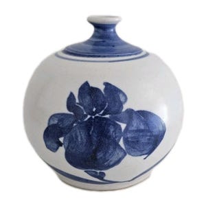 May include: A white ceramic vase with a round body and a narrow neck. The vase is decorated with a large blue floral design and blue accents around the neck. The vase is approximately 6 inches tall.