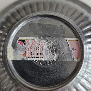 Puede incluir: Primer plano de la parte inferior de un cuenco peque&ntilde;o de metal plateado con borde acanalado. Una pegatina parcialmente rasgada est&aacute; adherida en el centro, con texto visible que incluye "21181" y "Maine". El cuenco tiene forma circular.