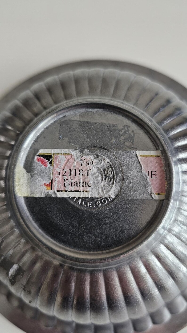 Puede incluir: Primer plano de la parte inferior de un cuenco peque&ntilde;o de metal plateado con borde acanalado. Una pegatina parcialmente rasgada est&aacute; adherida en el centro, con texto visible que incluye "21181" y "Maine". El cuenco tiene forma circular.